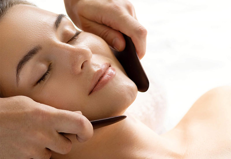 A person receiving a Gua Sha facial treatment, with a smooth jade stone tool gently scraping along the contours of the face, promoting relaxation and skincare benefits.